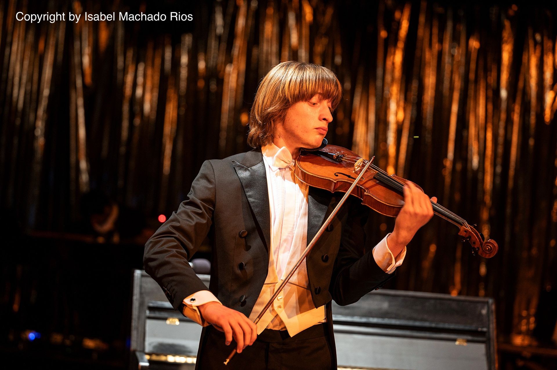 Person in formal attire playing a violin on stage with a dark, shimmering background.